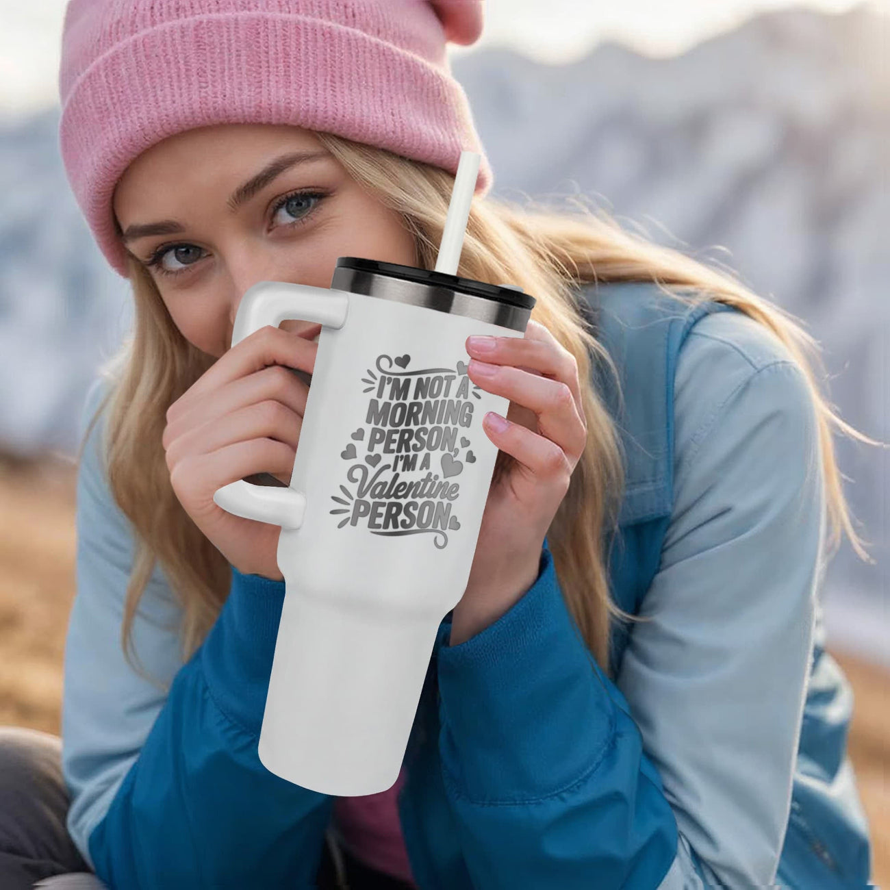 Person wearing a pink beanie and blue jacket holding a white tumbler with text outdoors.